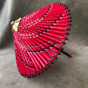May include: A red and black paper parasol with a decorative design. The parasol has a black handle and a gold-colored top. The edges are lined with small black beads. The parasol is approximately 10 inches in diameter.