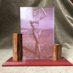 May include: A vintage photo frame with a sepia-toned image of a woman in a swimsuit. The frame has a red base and two wooden supports. The photo is of a woman on a beach.