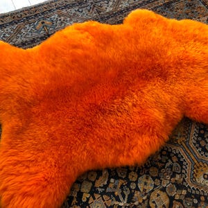 May include: A vibrant, solid orange sheepskin rug with a thick, plush, and shaggy texture. The rug features an irregular, natural pelt shape with soft, dense fibers that create a deep, fuzzy appearance across the entire surface of the item.