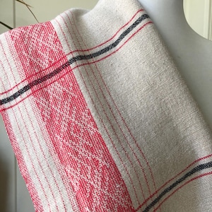 May include: A beige and red woven textile with a plaid pattern. The textile features red and white stripes and a red and white diamond pattern. Black stripes border the plaid design. The textile is draped over a white mannequin.