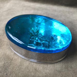 Op de afbeelding: Een klein ovaalvormig sieradendoosje met een zilveren basis en een helderblauw glazen deksel. Het glazen deksel is doorschijnend en heeft een geribbelde textuur.