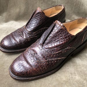 May include: A pair of brown leather shoes with a brogue design. The shoes have a textured, alligator-like pattern and a pointed toe.