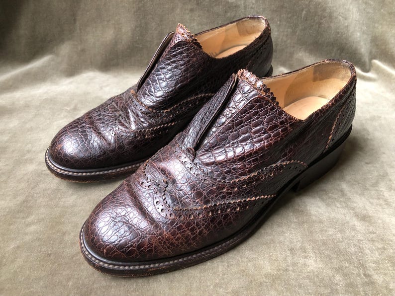 May include: A pair of brown leather shoes with a brogue design. The shoes have a textured, alligator-like pattern and a pointed toe.