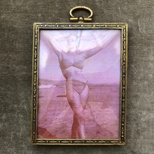 May include: Vintage brass picture frame with a photograph of a person in a swimsuit. The frame has an ornate design and a small loop at the top. The photograph has a sepia tone with a purple hue.
