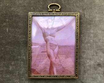 Può includere: Cornice vintage in ottone con una fotografia di una persona in costume da bagno. La cornice ha un design ornato e un piccolo anello in alto. La fotografia ha una tonalità seppia con una sfumatura viola.