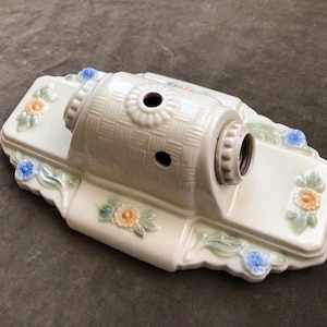 Puede incluir: Luminaria de cerámica blanquecina con textura trenzada en el cilindro central. La base rectangular presenta detalles florales en azul, naranja y verde. Tiene dos agujeros y un portalámparas.