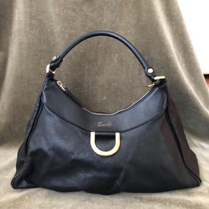 May include: Black leather handbag with a curved handle and gold-toned hardware. The bag features a top zipper closure and a large, open design. The brand name "Gucci" is visible on the front of the bag.