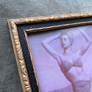 May include: Vintage framed photograph featuring a woman in a swimsuit. The frame is black with ornate gold detailing. The photograph has a sepia tone, with the woman posing with her arms above her head.