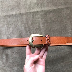 May include: A brown leather belt with a gold metal buckle and a braided leather detail.