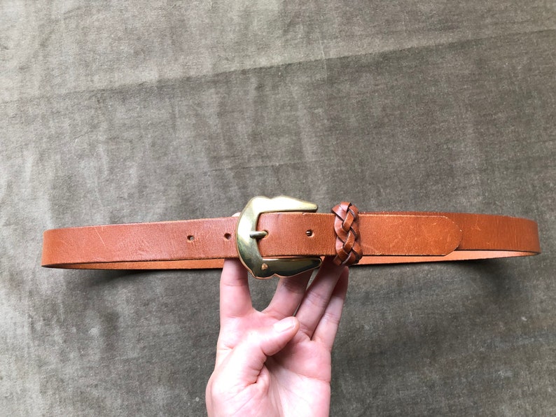 May include: A brown leather belt with a gold metal buckle and a braided leather detail.