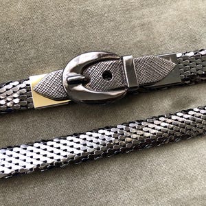 Könnte beinhalten: Ein silberner Metallgürtel mit einer anthrazitfarbenen Schnalle. Der Gürtel besteht aus kleinen, miteinander verbundenen Metallstücken, die einen Ketteneffekt erzeugen. Die Schnalle ist dunkelgrau mit einem strukturierten Design und einem goldfarbenen Akzent.