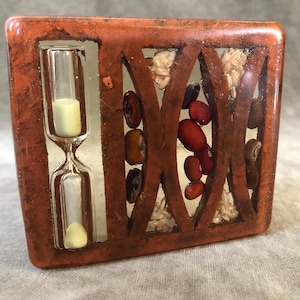 以下が含まれることがあります： 透明な砂時計が入った、小さな長方形の茶色の樹脂製の箱。箱には、赤と白の豆やその他の自然な要素で装飾されたデザインが施されています。