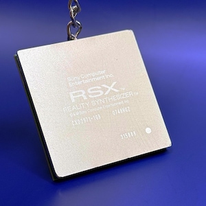 Puede incluir: Un llavero cuadrado de metal plateado con forma de chip de procesador informático. La superficie está grabada con el texto Sony Computer Entertainment Inc., RSX, REALITY SYNTHESIZER, CXD2971-1GB, 0748HGZ y 315888, unido a una cadena de metal plateado.