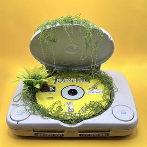 Può includere: Una console per videogiochi grigio chiaro con il coperchio aperto, contenente un disco di gioco Rayman giallo. La console è decorata con piccole foglie di felce artificiali verdi e rampicanti verdi che ricadono sul coperchio e sul disco.