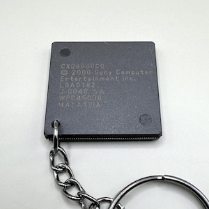 Puede incluir: Un llavero de componente electrónico negro cuadrado con el texto CXD8606CQ, 2000 Sony Computer Entertainment Inc., L9A0182, J 0046, WPC48038 y MALAYSIA. El componente está unido a una cadena de metal plateado y una argolla circular.