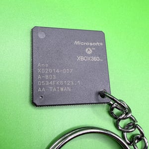 Puede incluir: Un llavero cuadrado de chip de computadora negro con el logotipo de Microsoft Xbox 360 y el texto X02014-007, A-B03, 0534FK6123.1 y AA TAIWAN. El componente está unido a una anilla de metal plateado mediante una cadena corta, mostrando marcas técnicas.