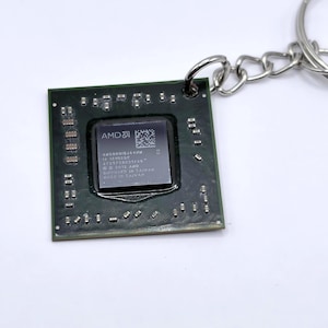 Pode incluir: Um porta-chaves de chip de processador de computador quadrado verde-escuro com uma placa metálica prateada central com o logótipo AMD e texto incluindo AM5000IBJ44HM, 2012 AMD e Made in Taiwan. O chip tem pequenos pontos de contacto prateados.