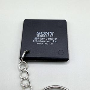 Puede incluir: Un llavero de componente electrónico cuadrado negro con texto impreso en blanco que dice SONY CXD8561Q 1995 Sony Computer Entertainment Inc. KOREA 9631EBI. El componente está unido a una cadena de metal de color plateado y una anilla circular.