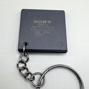 Puede incluir: Un llavero cuadrado de chip de procesador de computadora gris oscuro con una cadena y un anillo de metal plateado. El chip tiene impreso el texto SONY, CXD8561CQ, 1996 Sony Computer Entertainment Inc., KOREA, 9934EB1 y F5629ZA1 en gris claro.