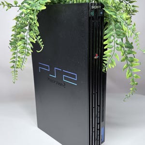 Könnte beinhalten: Eine schwarze Sony PlayStation 2 Spielkonsole, die vertikal aufgestellt ist. Die Vorderseite zeigt das blaue PS2-Logo und den Schriftzug PlayStation 2. Die Konsole verfügt über einen Ein-/Ausschalter, eine Reset-Taste, eine Auswurftaste und Steckplätze.