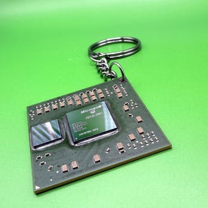 Puede incluir: Un llavero cuadrado que presenta un chip de procesador de computadora de color verde oscuro con acabado metálico. El chip central tiene impreso el texto Microsoft XBOX 360 y X819195-002. Incluye una anilla de metal oscuro unida por una cadena.