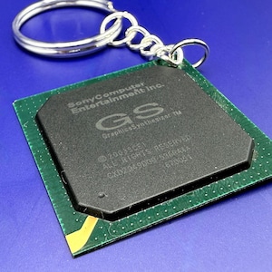 May include: A square green computer chip keyring featuring a black processor component with the text Sony Computer Entertainment Inc. GS Graphics Synthesizer, 2003 SCEI, ALL RIGHTS RESERVED, and CXD29480GB 536R4AA 670451 printed on the surface.