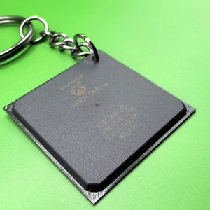 Puede incluir: Un llavero cuadrado de color gris oscuro con el logotipo de Xbox One y la marca Microsoft. La superficie incluye texto técnico grabado y un pequeño orificio en la esquina superior, unido a una anilla de metal plateado mediante una cadena corta.
