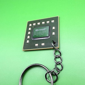 Puede incluir: Un llavero de procesador de computadora cuadrado de color verde con una cadena y un anillo de metal oscuro. La superficie central del chip muestra el texto Microsoft, XCPU C-A01, X812480-004, 0805590FC y C TAIWAN. Tiene contactos dorados en los bordes.