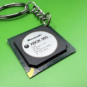 Puede incluir: Un llavero cuadrado que presenta un componente electrónico negro con un centro plateado circular. La etiqueta plateada muestra el texto Microsoft Xbox 360, XSB, X02047-027, B-R0, 0818PTD1894 y A Taiwan. Se incluye una anilla y cadena de metal.