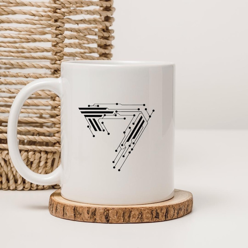 May include: White ceramic mug with a black circuit board design in the shape of the number seven. The mug sits on a round wooden coaster. A woven basket is in the background.