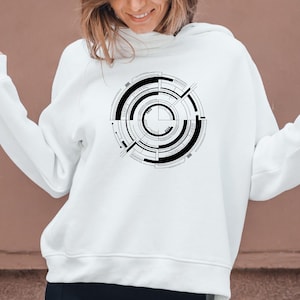 May include: White hoodie featuring a black and white circular geometric design on the front. The design consists of concentric circles, lines, and rectangular shapes. The hoodie has a drawstring hood and long sleeves.