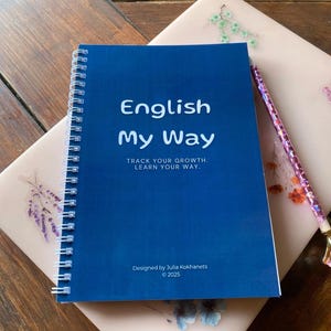 May include: A spiral-bound notebook with a navy blue cover, titled "English My Way." The cover also includes the text "Track Your Growth. Learn Your Way." and the designer's name. The notebook rests on a light pink surface with floral accents, next to a decorative pen.