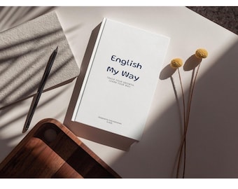 Puede incluir: Un libro blanco titulado "English My Way" con texto azul, junto a un cuaderno gris, un lápiz negro, una bandeja de madera y flores amarillas secas. La portada del libro incluye el lema "Track your growth, learn your way."