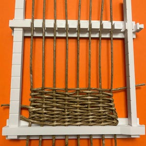 May include: A white rectangular weaving loom with vertical brown reeds and woven brown material. The loom is set against an orange background. The weaving process is partially complete.