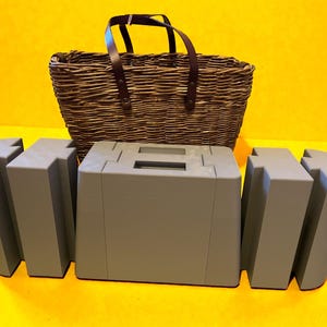 May include: A wicker picnic basket with a brown leather handle is positioned behind three grey, geometric, modular storage containers. The containers are in front of the basket on a yellow surface.