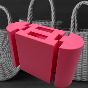 Könnte beinhalten: Eine leuchtend rosa geometrische Skulptur mit einer rechteckigen Mittelform und zwei parallelen rechteckigen Öffnungen. Zwei gebogene, zylindrische Teile flankieren die Mittelform. Der Hintergrund zeigt zwei geflochtene Körbe.
