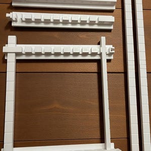 Könnte beinhalten: Weiße Kunststoff-Regalteile, darunter zwei vertikale Stützen, ein rechteckiger Rahmen und zwei horizontale Teile mit gekerbten Kanten. Die Teile sind auf einer braunen Holzoberfläche angeordnet.