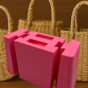 May include: A bright pink, geometric sculpture with a rectangular form and cut-out sections. Woven wicker handbags with arched handles are in the background. The sculpture is the focal point, set against a warm, neutral backdrop.