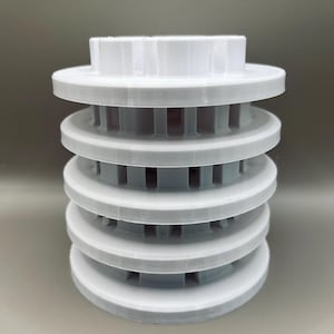 Op de afbeelding: Een stapel van vijf lichtgrijze, cirkelvormige objecten met een witte bovenkant. Elke cirkelvormige laag heeft een platte, schijfvormige vorm met een reeks verticale ribbels langs de binnenrand. De bovenkant is wit en heeft een bloemachtig ontwerp.