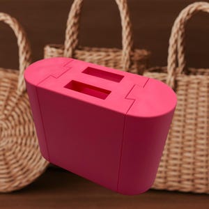 Könnte beinhalten: Ein leuchtend pinkfarbener Toaster mit zwei Schlitzen. Der Toaster hat einen rechteckigen Körper mit abgerundeten Kanten und einer glatten Oberfläche. Geflochtene Korbtaschen sind im Hintergrund.