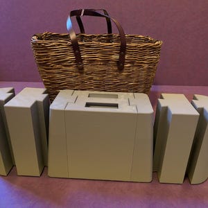 Könnte beinhalten: Ein geflochtener Picknickkorb mit braunem Ledergriff steht hinter drei hellbeige geometrischen, modularen Teilen. Die Teile scheinen so konzipiert zu sein, dass sie zusammenpassen. Der Korb ist leer.