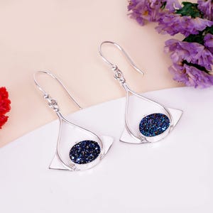 May include: A pair of silver drop earrings. Each earring has a teardrop-shaped frame with a square base and a dark blue, glittery oval centre. The earrings are displayed on a white surface with purple flowers.