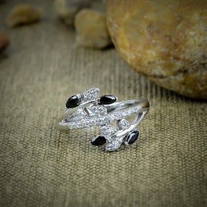May include: A silver ring with a leaf design, adorned with black and clear gemstones. The ring has a split shank and is displayed against a neutral backdrop, highlighting the jewellery's intricate details.
