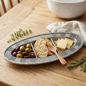 May include: Oval, divided serving tray with olives, crackers, and cheese. The silver-colored tray has a geometric design around the edges. A wooden fork rests on the tray. The tray is on a wooden table with a white bowl in the background.