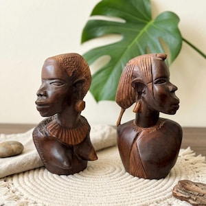 May include: Two dark brown wooden busts of African women. The busts are carved with detailed features, including hairstyles, earrings, and necklaces. The busts are displayed on a cream-colored macrame placemat.