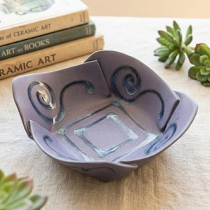 May include: A square, lavender ceramic bowl with a unique, folded rim design. The bowl features a central diamond pattern and swirling blue accents. The bowl is displayed on a neutral surface, with books and greenery in the background.