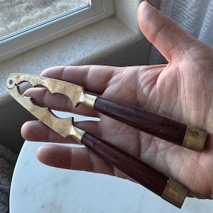 May include: Vintage nutcracker with brass jaws and dark wood handles. The nutcracker is held in a hand, showcasing its design. The brass jaws have a unique, decorative shape. The handles are a rich, dark brown color, with brass accents.
