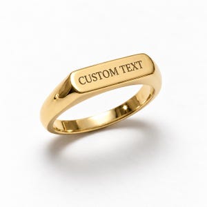 Puede incluir: Un anillo de sello en tono dorado pulido con una superficie superior plana y rectangular. La parte superior plana está grabada con las palabras CUSTOM TEXT en una fuente serif oscura y clara. La banda es redondeada y tiene un acabado dorado.