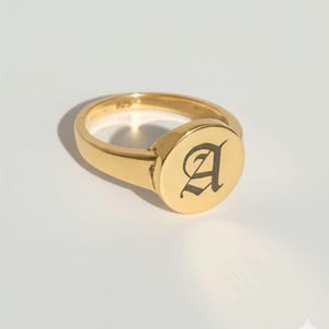 以下が含まれることがあります： 平らで円形の面を持つ金色のシグネットリング。リングの表面には、エレガントで様式化されたフォントで黒い文字「A」が描かれています。リングは光沢のある研磨された金属でできており、バンドは滑らかで丸みを帯びています。