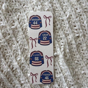 May include: A rectangular bookmark featuring four miniature hockey jerseys in navy blue, with white and brown accents. Each jersey displays a name and number: Graham 44, Logan 22, Di Laurentis 66, and Tucker 46. Burgundy bows are placed between each jersey.